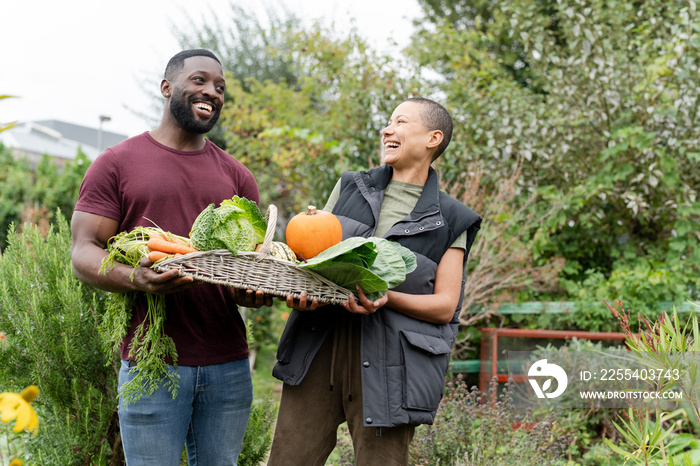 Portrait of smiling couple holding basket with fresh vegetables in urban garden