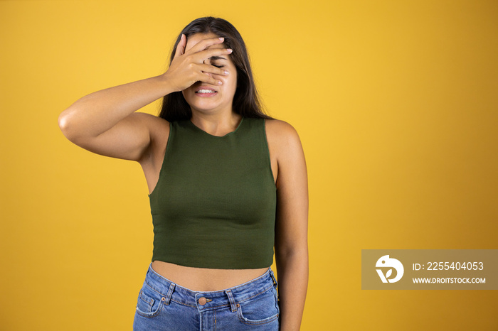 Young beautiful woman standing over isolated yellow background peeking in shock covering face and eyes with hand, looking through fingers with embarrassed expression