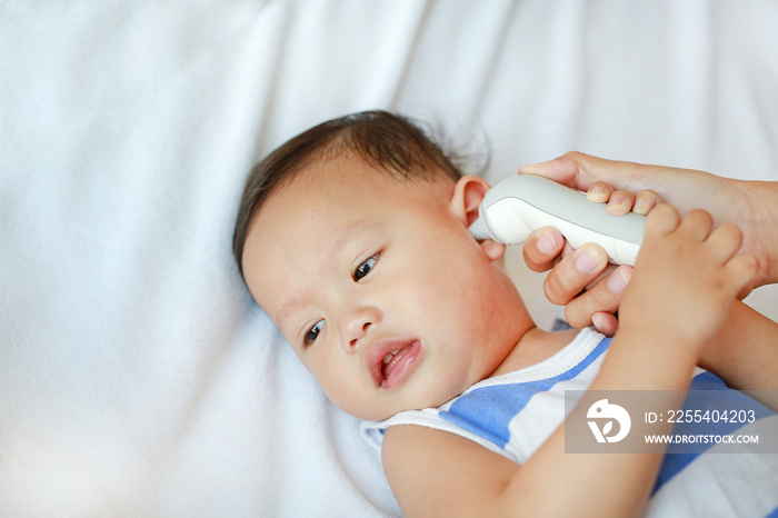 Mother takes temperature for baby boy with ear thermometer on bed at home.