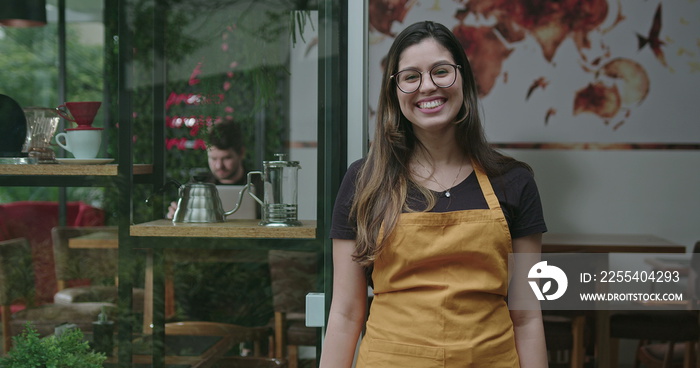 One happy female employee standing in front of small business coffee shop smiling. Young woman barista employed at a cafe restaurant portrait face