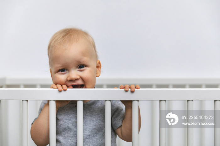 smiling toddler standing in the bed and holds by the cot rails. looking at camera