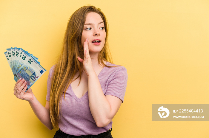 Young caucasian woman holding banknotes isolated on yellow background is saying a secret hot braking news and looking aside