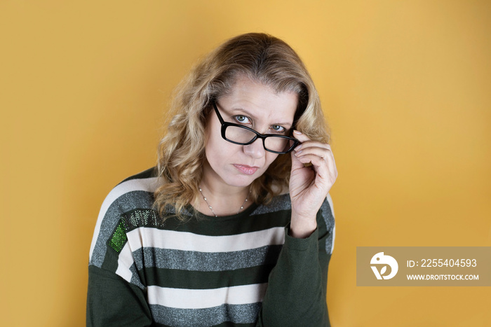 Mature woman with a stern sad look in glasses, a woman looking out from under glasses on a yellow background, a woman’s eyesight has deteriorated and she must wear glasses