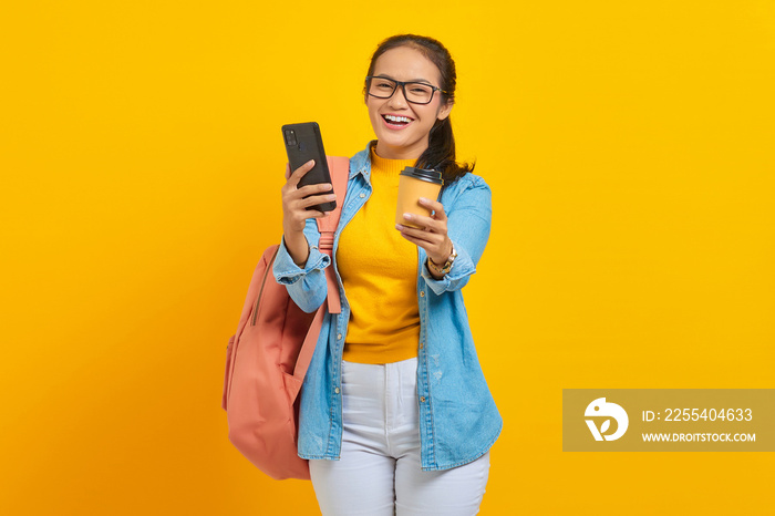 Portrait of cheerful young Asian woman student in denim clothes with backpack holding cup of coffee and smartphone isolated on yellow background