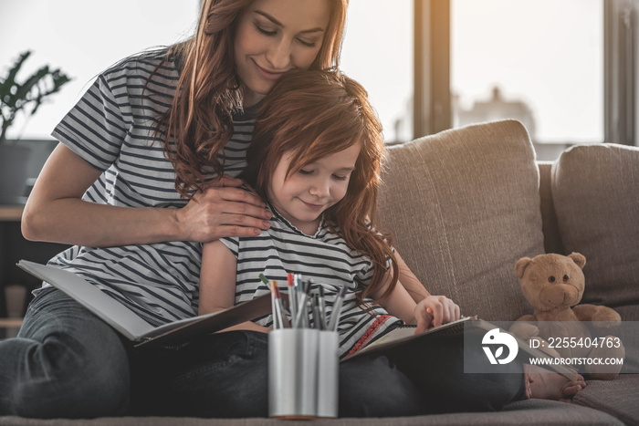 Time together. Full length portrait of thoughtful mother teaching her daughter how to draw. They are sitting bonding to each other on couch at home, holding sketchbooks and crayons