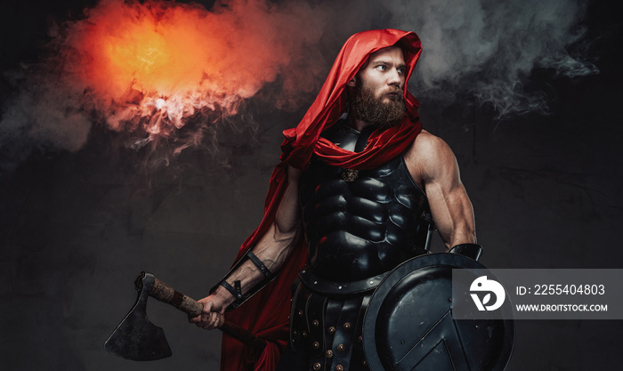 Armoured and bearded rome warrior in red cloak and black armor posing holding axe and shield in dark room with steam.