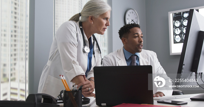 Portrait of millennial black doctor using computer while senior colleague directs him. Two doctors working inside medical office looking at computer monitor and typing