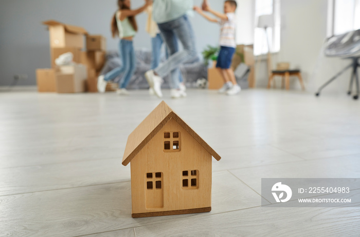 Buying house for family. Small model of wooden house on floor in living room on background of family that is happy to move. Buying house or apartment concept. Selective focus.