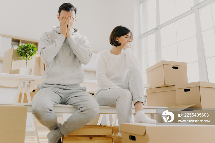 Worried couple pose near stack of cardboard boxes, have to move in other flat, have no money for rent, have dejected sad expressions, sit together in white room, have family conflict and divorce