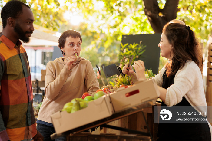 Multiracial couple tasting bio organic produce at farmers market, selling locally grown fruits and vegetables at farmers market. Young family tasting natural eco produce, food sampling.