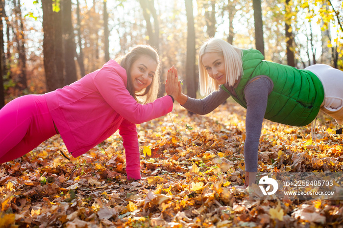 Two active fit women practice plank exercise and high five in autumn forest. Outdoor workout