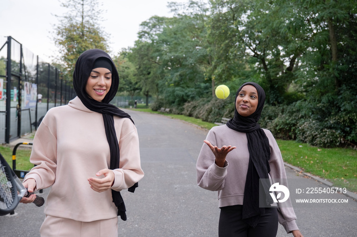 Two women in hijabs walking in park with tennis racket and ball