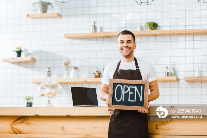 handsome cashier in brown apron standing near wooden bar counter and holding chalkboard with open lettering in coffee house