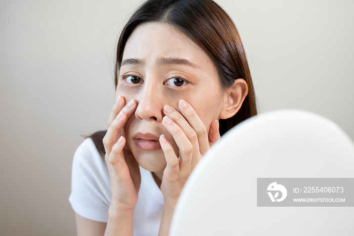 Sleepless Asian woman looking her face in the mirror and worry about dark circles under eyes