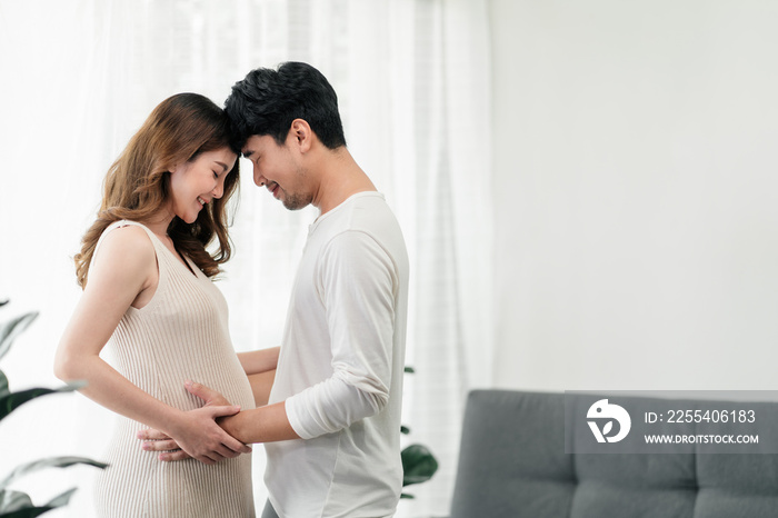 A happy asian couple. A beautiful pregnant wife and her  handsome husband are smiling happily with their heads touched and their hands grabbing the wife’s tummy to feel their baby.