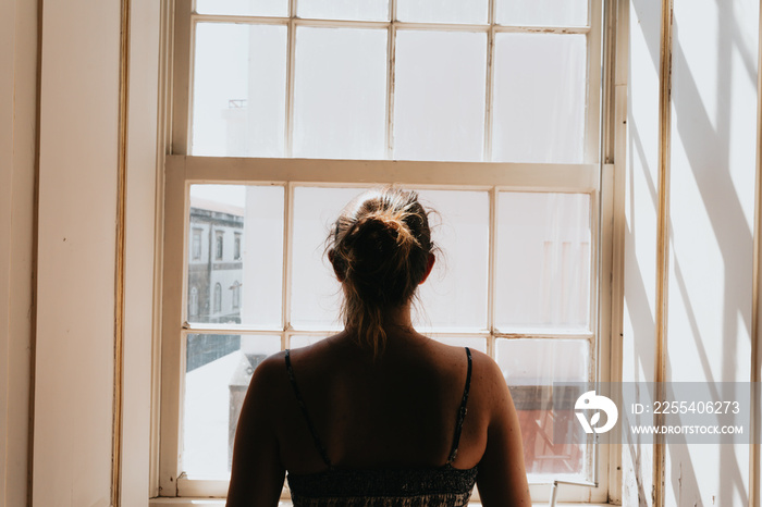 Young woman using a dress looking through a window, giving the back to the camera, stress and anxiety concepts, dark image, sadness