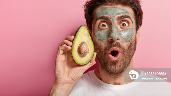 Spa and beauty concept. Cropped image of handsome man stares with widely opened eyes at camera, applies natural clay mask on face, holds slice of avocado, visits spa salon, isolated on pink wall