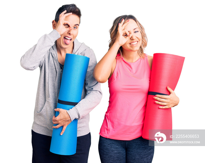 Couple of women holding yoga mat smiling happy doing ok sign with hand on eye looking through fingers