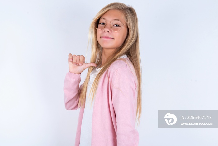 Closeup of cheerful caucasian blonde little girl wearing pink jacket and glasses over white background looks joyful, satisfied and confident, points at himself with thumb.