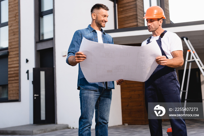 Workman in helmet holding blueprint near man and building outdoors