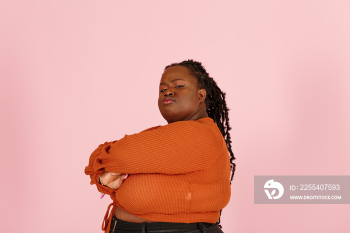 Critical young black plus size body positive woman with dreadlocks in orange top looks at camera crossing arms on pink background in studio