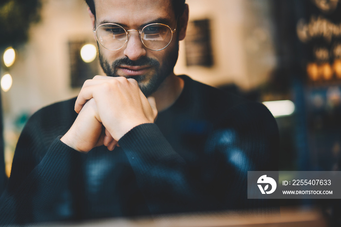 Cropped image of thoughtful freelancer in trendy optical glasses reading trade news on computer. Concentrated male economist searching information on laptop computer while sitting in cozy interior