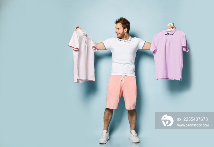 Young handsome man in pink shorts and white t-shirt standing decide, holding two polo shirts feeling crazy unable to chose