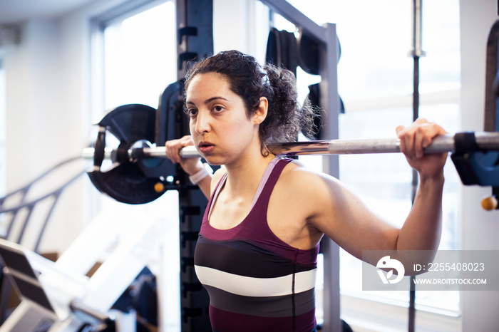 Confident female athlete holding barbell looking away while standing in gym