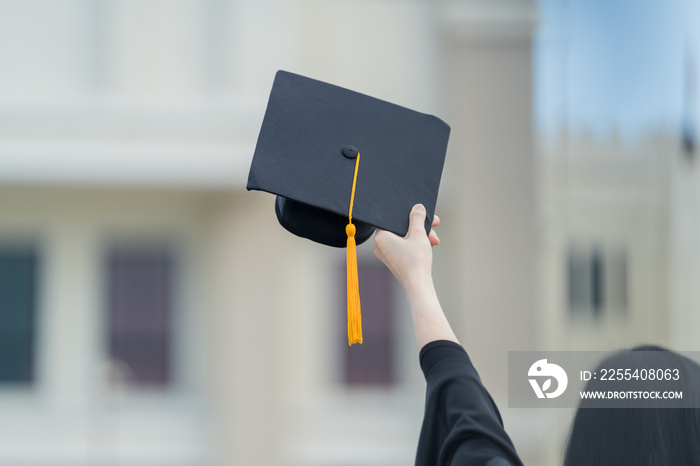 A young beautiful Asian woman university graduate in graduation gown and mortarboard holds a degree certificate stands in front of the university building after participating in college commencement