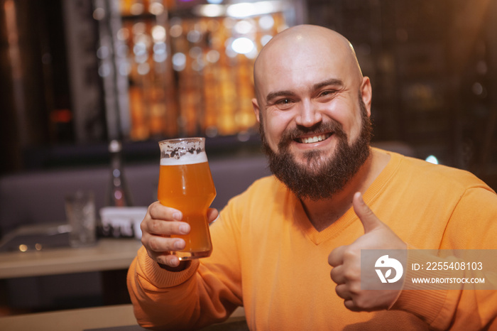 Excited bearded bald man smiling to the camera, showing thumbs up while having a glass of delicious beer at the pub