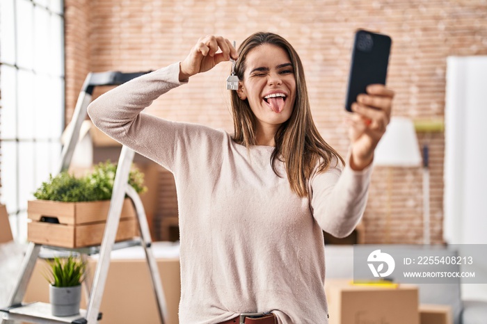 Beautiful woman holding keys of new home showing on video call sticking tongue out happy with funny expression.