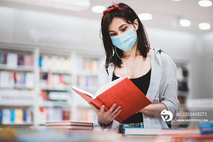 Young female student dressed in trendy outfit preparing for exam and reading a book at the library.Cute hipster girl with face medical mask looking for study references at University during pandemic