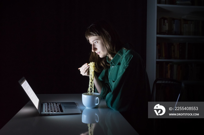 Young female at laptop computer eats instant ramen noodles late in the evening. Woman working or studying online overtime at night has fast dinner - concept of unhealthy junk food at workplace