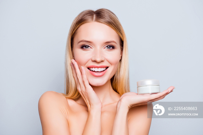 Close up portrait of happy gorgeous smiling attractive satisfied young woman with beaming smile, she is holding a cream jar and touching her cheek, isolated on grey background