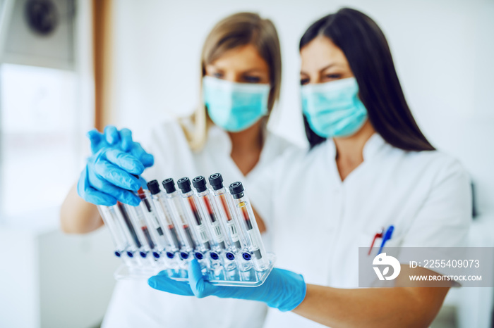 Two dedicated caucasian lab assistants in uniforms, with masks and rubber gloves standing in lab and looking at test tubes with blood samples.