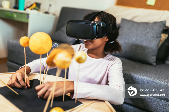 Happy african american school girl making a solar system for a school science project at home using VR glasses to immerse herself in metaverse