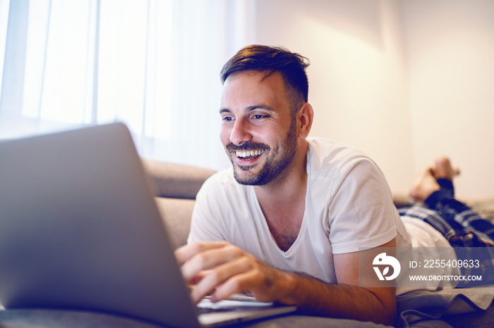 Joyful caucasian man in pajamas lying on stomach on sofa in living room and surfing on internet.