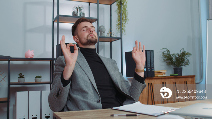 Businessman in suit working on laptop computer, meditating, doing yoga breathing exercise in lotus position at home office desk. Calm serene freelancer man taking break. Busy occupation. Peace of mind