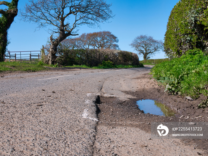 Surface damage to tarmac on a rural road is marked for repair with white spray paint. Taken on Wirral in the UK on a sunny day in spring.