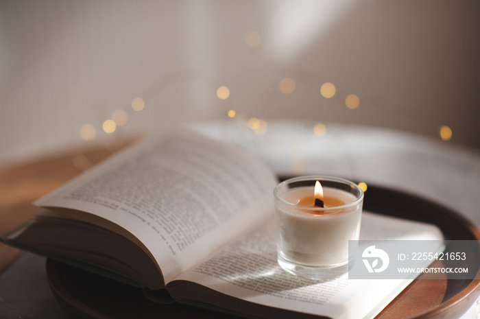 Burning scented candle staying on open paper book on wooden tray over glow lights on table in bedroom. Cozy home atmosphere.