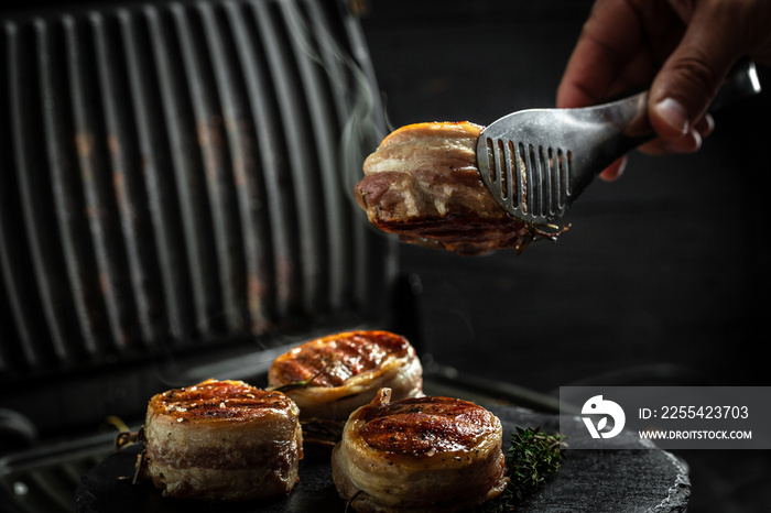 Medallions steaks from the beef tenderloin covered bacon on grill Dark background. Cooking beef steak on grill by chef hands