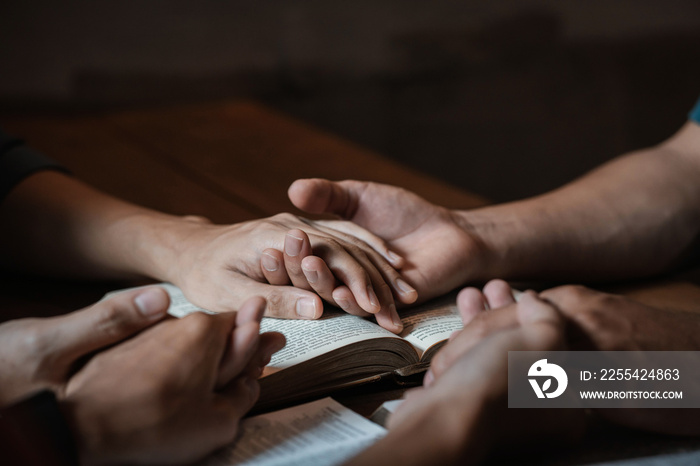 A group of young Christians holding hands in prayer for faith and scriptures on a wooden table as they pray to God.