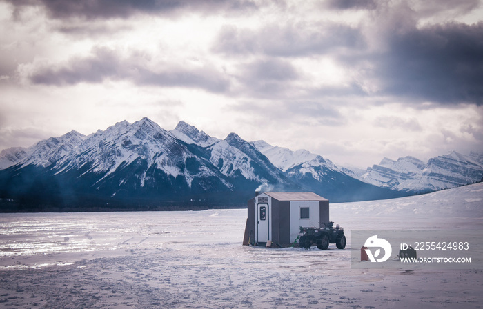 Ice shack sitting on Lake Abraham in Alberta with a quad and the peaceful mountains in the distance