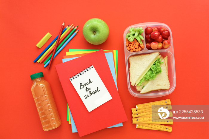 School supplies and lunch box with sandwiches, pieces of cucumbers and carrots, apricot, cherries, apple, bottle of juice on red background, back to school concept.