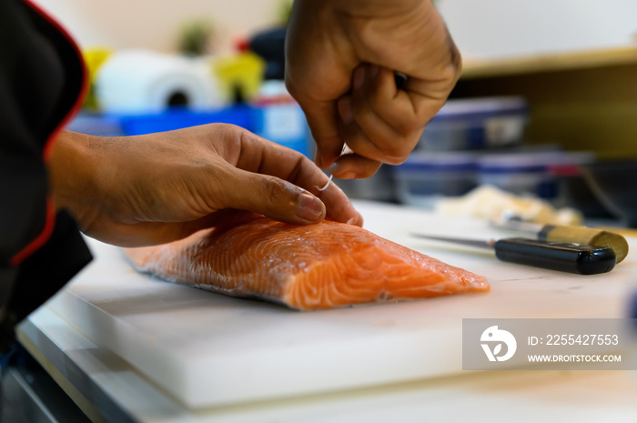 Fresh salmon fillet close-up on white cutting board table while fishbone removal with stainless steel tweezers ​by chef in cooking room. Selection focus on salmon fillet with Soft focusing shooting
