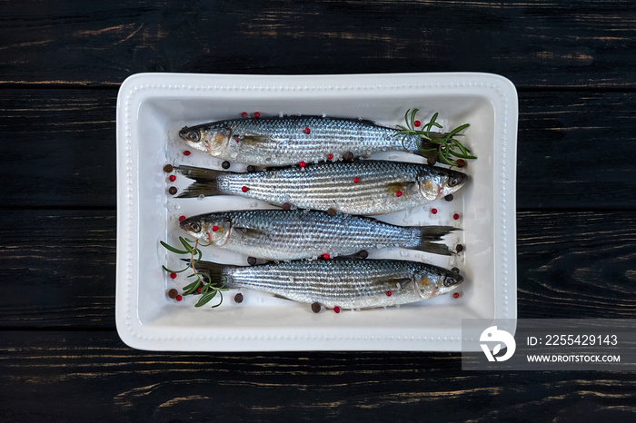 Raw mullet fish with spices and herbs ingredients in white ceramic backing dish on black wooden table ready for cooking. Cooking concept.