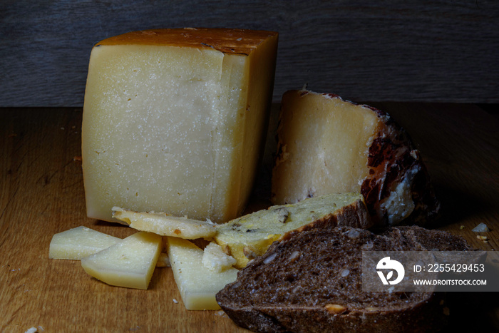 two pieces, triangles, portions of cheese, idiazabal and from the Pyrenees, one vertical and the other horizontal, with smaller portions of and slices of cereal and corn bread on a wooden table.