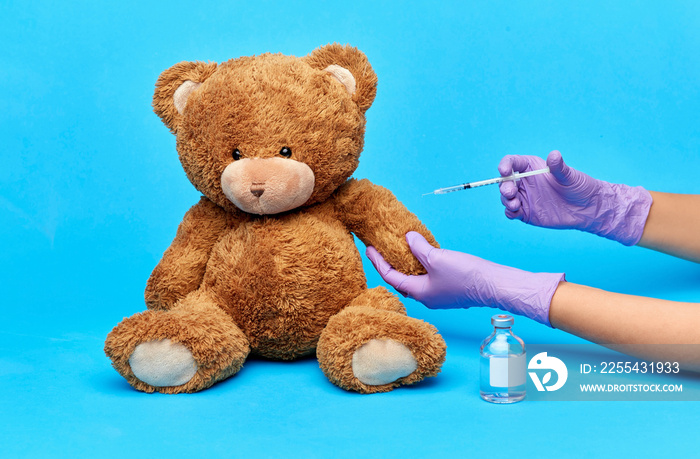 vaccination, healthcare and pandemic concept - hands of doctor in medical gloves with vaccine and syringe making injection teddy bear toy over blue background