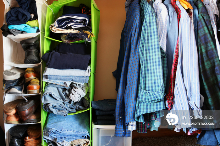 Private house closet with well organized casual man clothes.Man wardrobe with variety of shirts hung on hangers, shoes and pile of jumper, jeans and pants lain in a storage organizers pocket shelves.