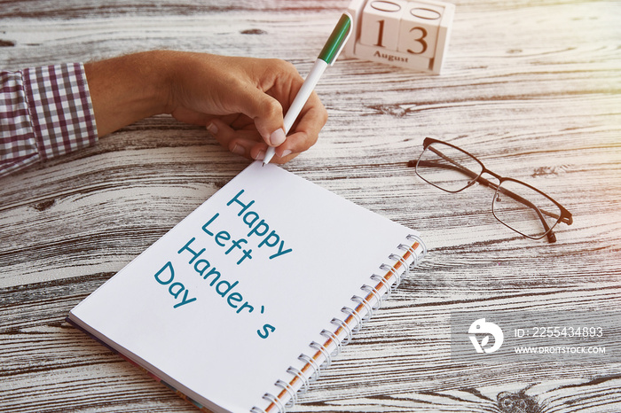 Man uses his left hand to wright in a notebook. Glasess and calendar with 13th of August - Day of Left Handers. Conceptual photo of lefties day. Copy space.